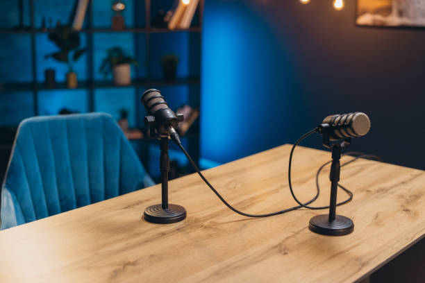 A wooden table set with two professional podcast microphones on stands, a plush blue chair, and a bookshelf in a dimly lit background with blue ambient lighting.